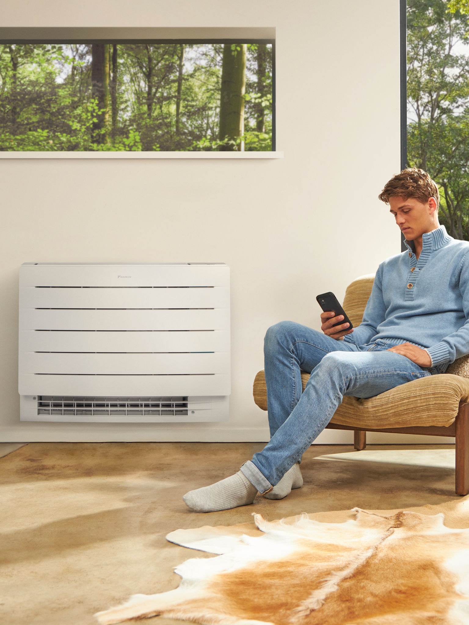 A man sitting in a light room looking at his phone with a heat pump on the wall behind him