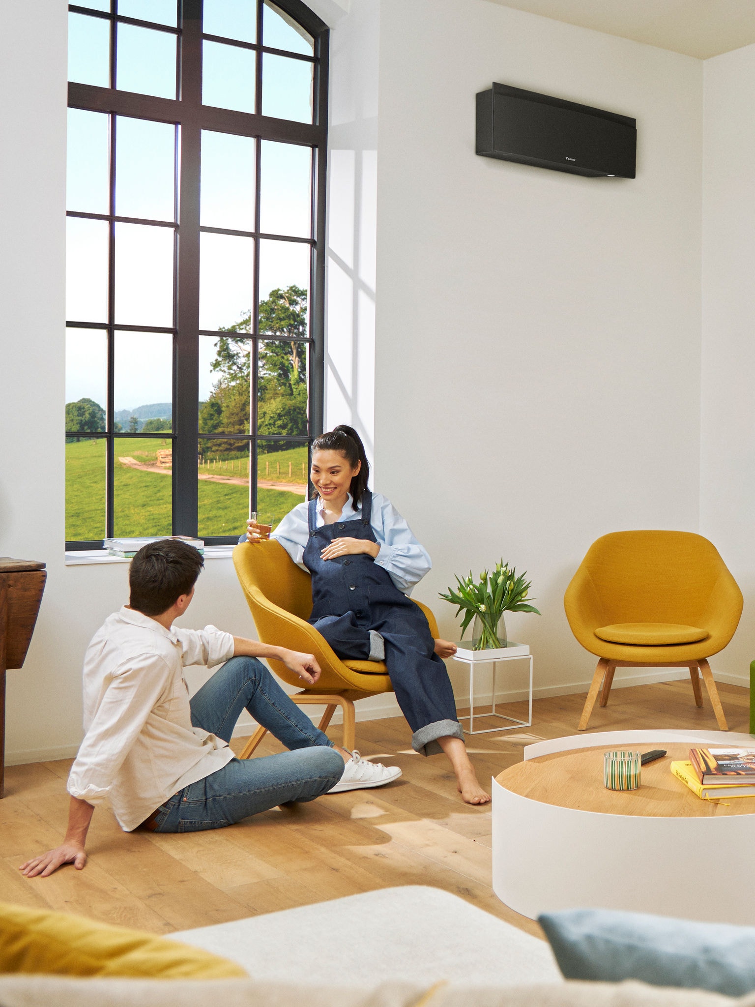 A man and a woman sitting in a bright room in front of large window with the sun shining outside and a black wall-mounted heat pump on the wall behind them