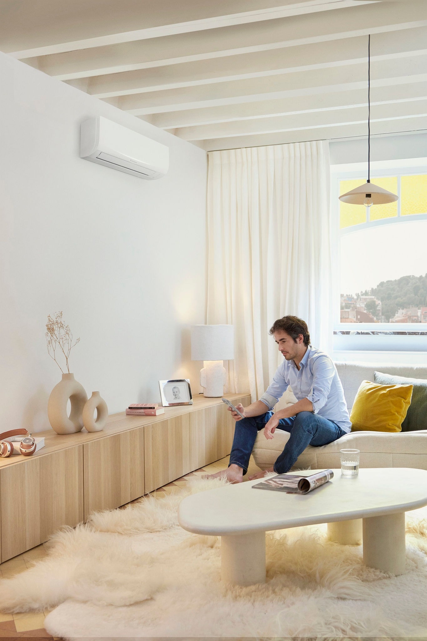 Man on phone sitting on couch under window Daikin Sensira White on wall left