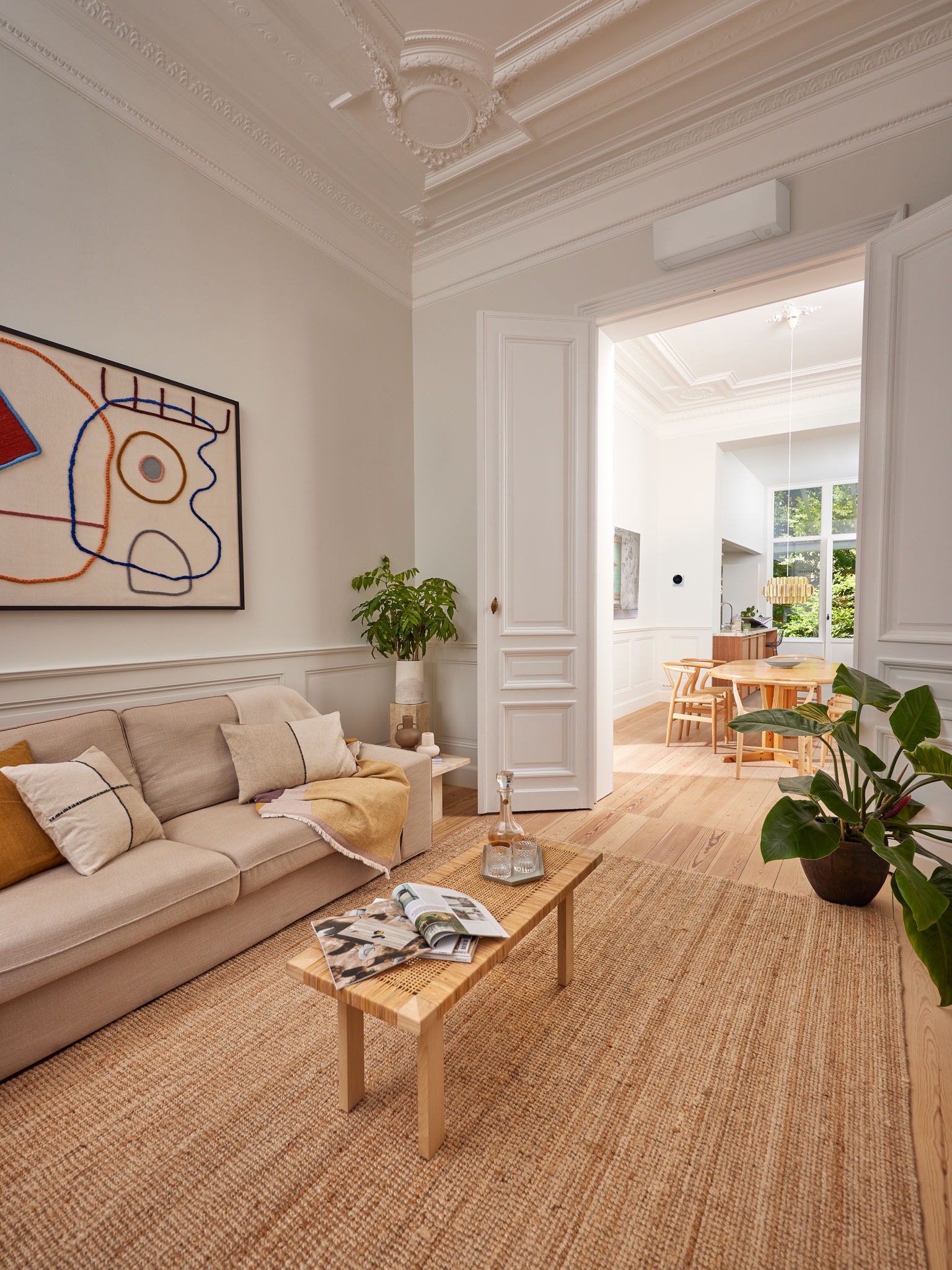 A white, wall mounted heat pump above a doorway leading from a bright sitting room with white walls and a cream sofa into a dining room with a table in front of a large window
